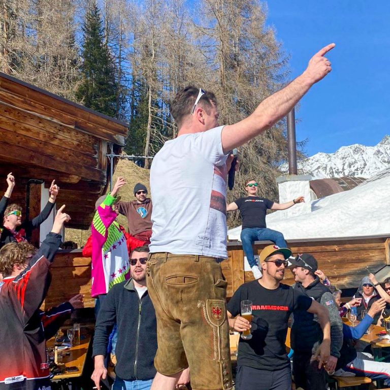 Menschen feiern fröhlich draußen in den Bergen, einige tanzen auf einer Terrasse.