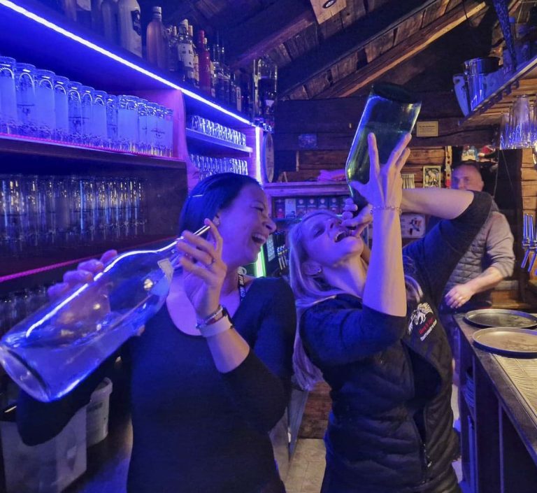 Zwei Frauen in einer Bar, die aus großen Flaschen trinken und lachen.